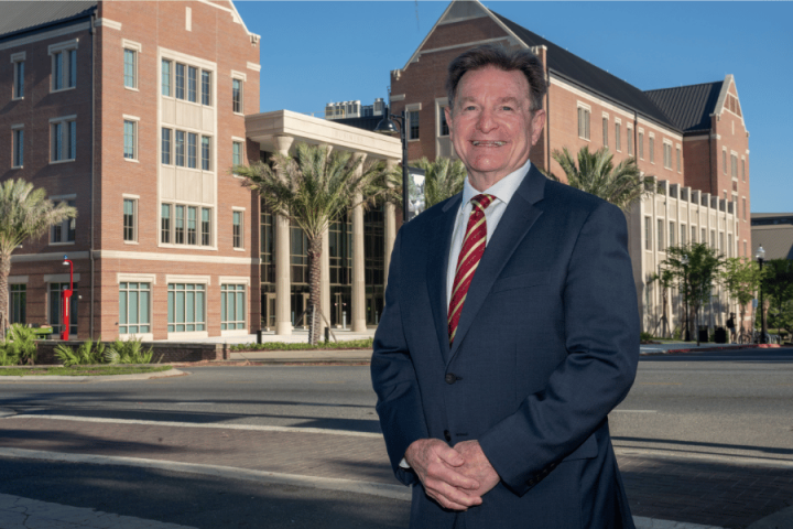 A smiling man in a navy blue suit and a red striped tie stands outdoors, with large brick university buildings and palm trees in the background.