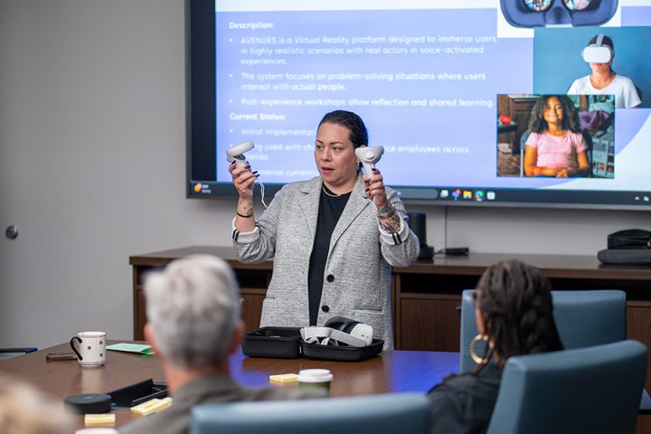 A woman in a grey blazer stands in a conference room demonstrating virtual reality equipment, holding up two VR controllers in front of a presentation screen.