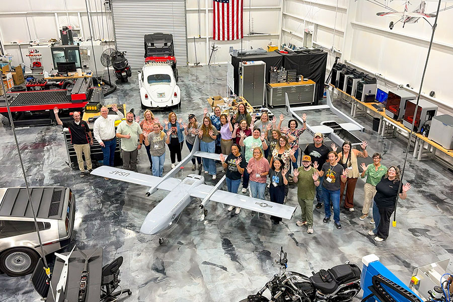 A high-angle photo shows a group of over 20 people waving inside a large workshop hangar as they gather around a grey military drone with USAF markings. The surrounding space features various vehicles, including a DeLorean and a white Volkswagen Beetle, alongside workbenches, 3D printers, and an American flag hanging on the back wall.